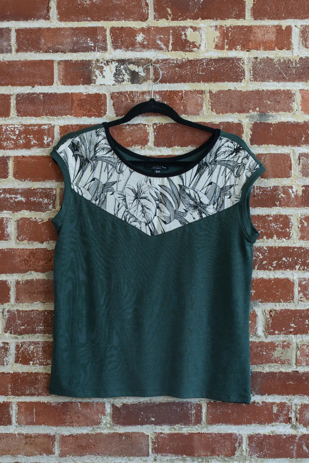 The Pepita Top from Marie C, a reversible t-shirt with a printed fabric yoke on one side, in Green, is shown hanging on a brick wall.