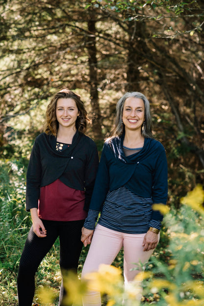 Two women standing side to side in a forest, wearing the Boreal Bolero by Marie C. One is wearing the Black and one is wearing the Navy. The bolero features contrasting fabric at the back, a high collar that can be folded down, and a single button closure. 