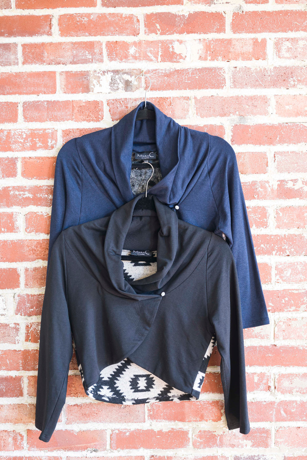 The Boreal Bolero by Marie C in Black  and Navy are shown hanging against a brick wall. The boleros feature contrasting fabric at the back, a high collar that can be folded down, and a single button closure. 