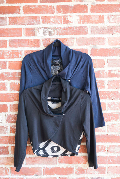 The Boreal Bolero by Marie C in Black  and Navy are shown hanging against a brick wall. The boleros feature contrasting fabric at the back, a high collar that can be folded down, and a single button closure. 