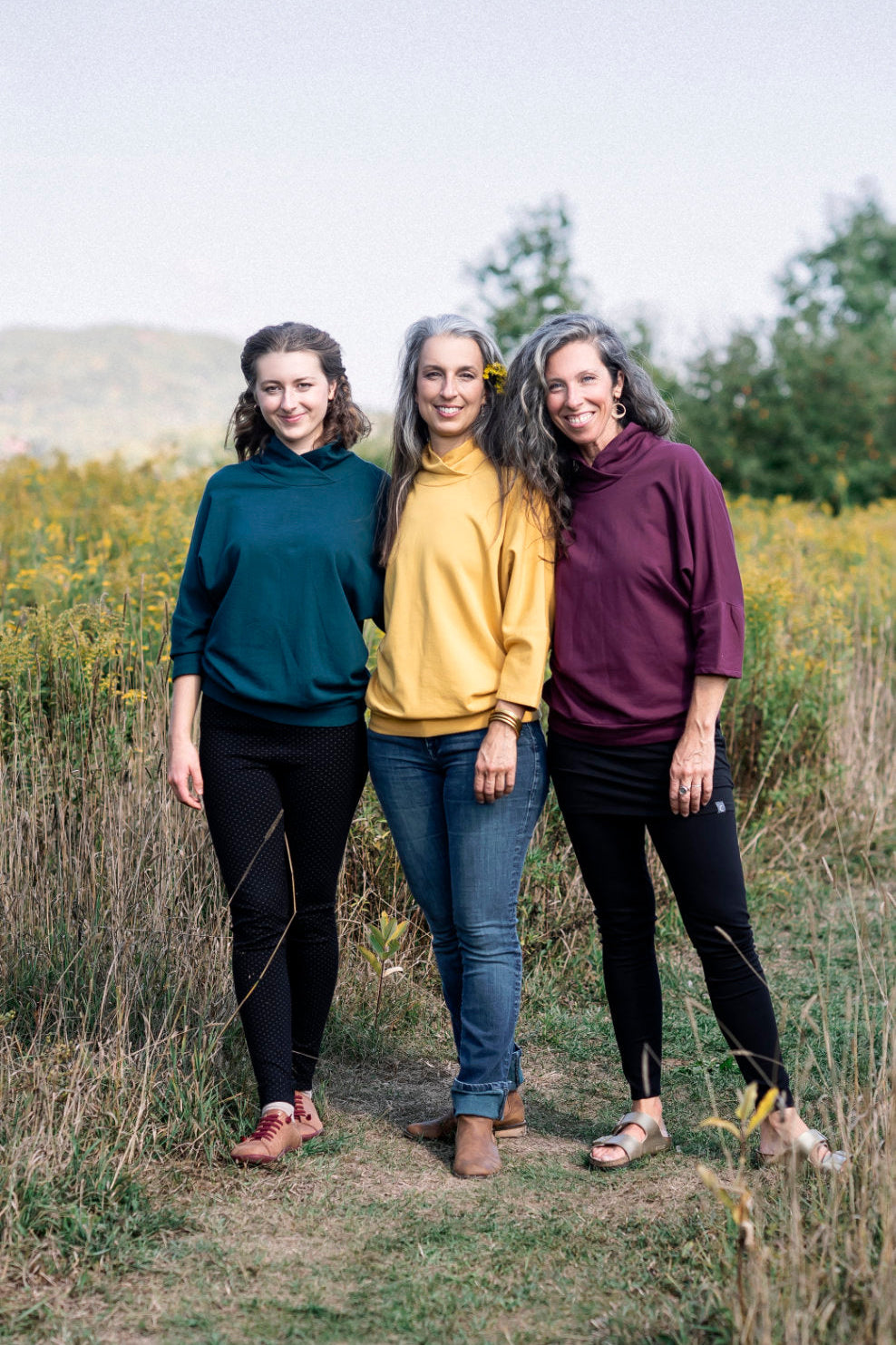 Three women wearing the BeAAA Sweater by Marie C in Everglade, Yellow and Wine, standing in a field 