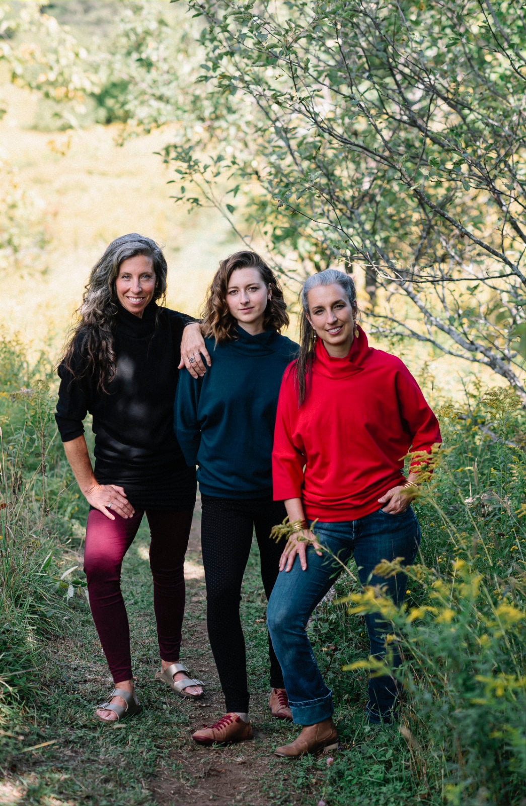 Three women wearing the BeAAA Sweater by Marie C in Everglade, Black, and Red, standing in a forest.