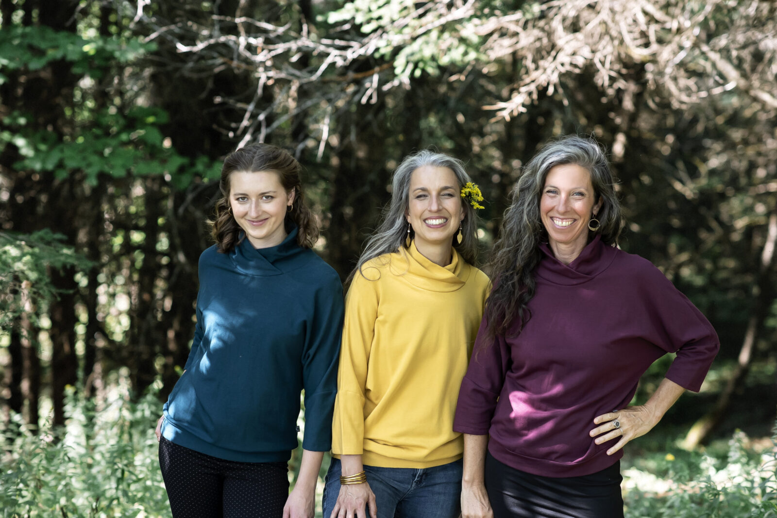 Three women wearing the BeAAA Sweater by Marie C in Everglade,Yellow, and Wine, standing in a forest. 