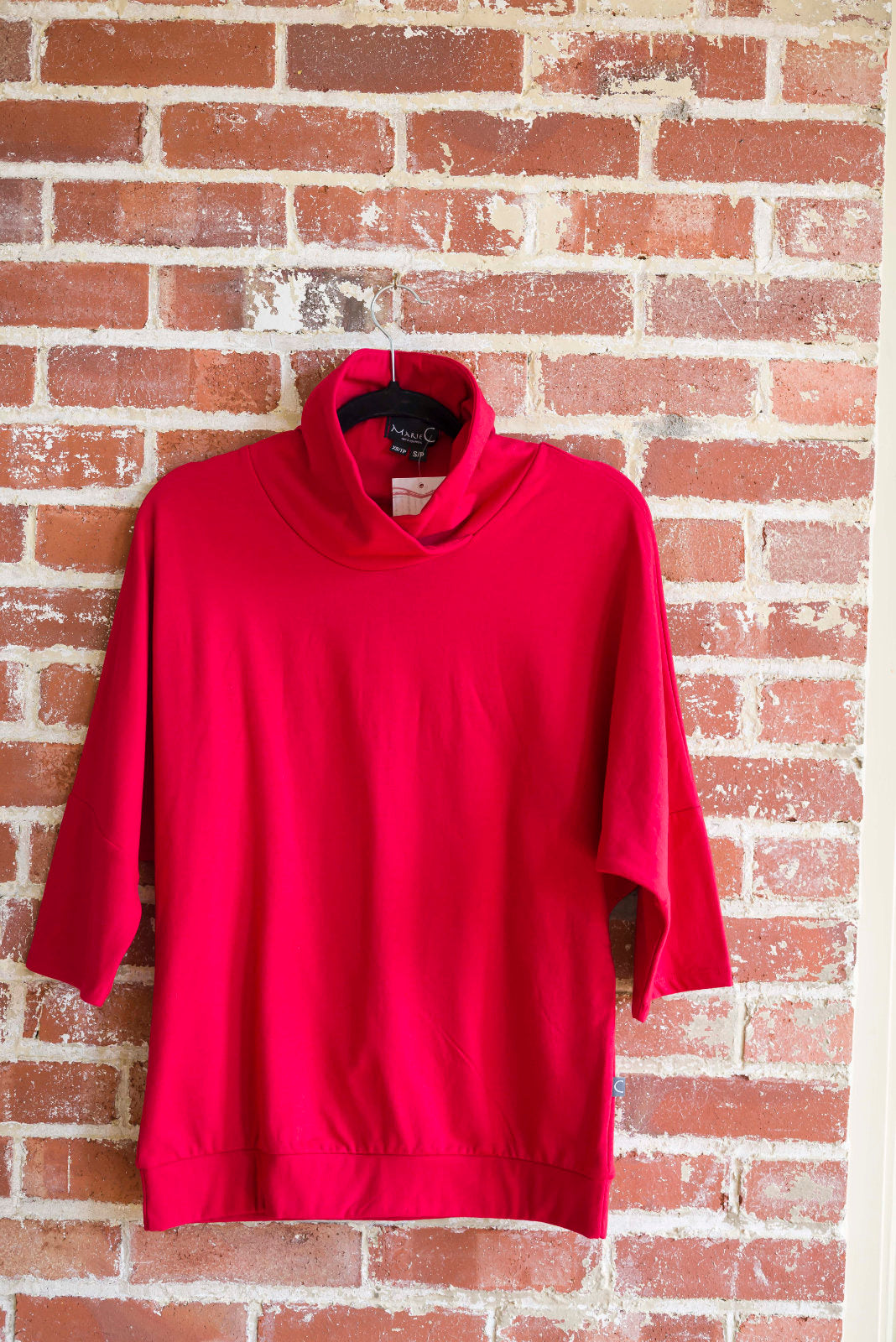 The BeAAA sweater by Marie C in Red on a hanger against a brick wall