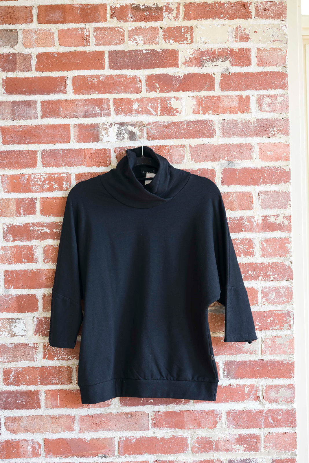 The BeAAA sweater by Marie C in Black on a hanger against a brick wall