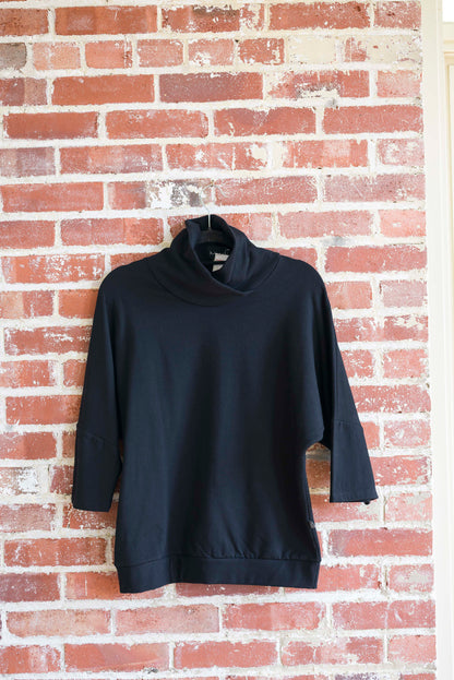 The BeAAA sweater by Marie C in Black on a hanger against a brick wall