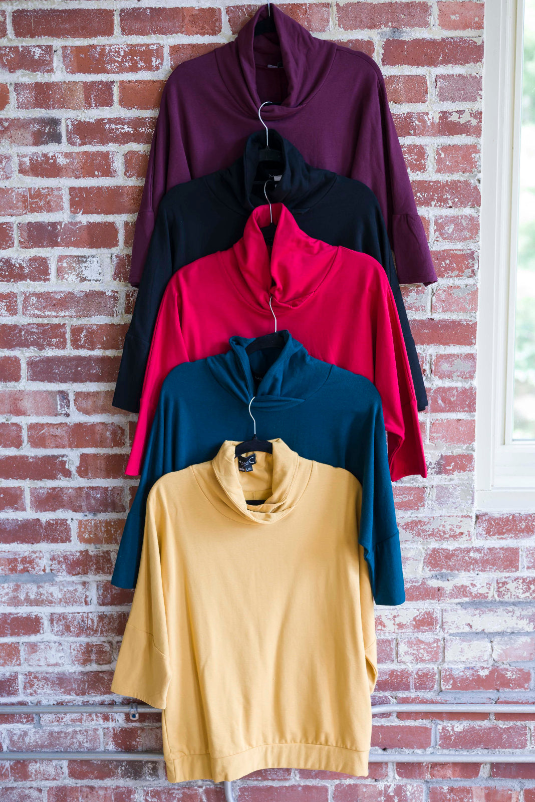BeAAA Sweaters by Marie C in Wine, Black, Red, Everglade , and Yellow are shown hanging against a brick wall. 
