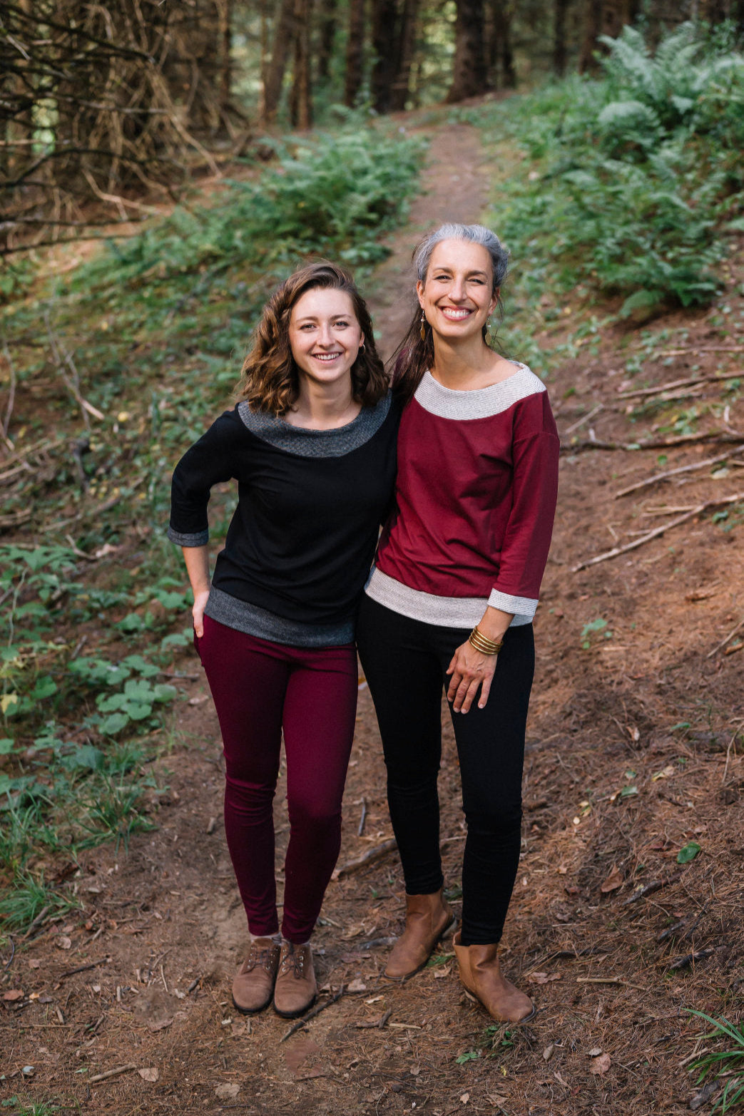 Two women wearing the Jaja Top by Marie C, one in Black and one in Wine. It's a sweatshirt with contrasting fabric bands at the wide neck, 7/8 sleeves, and hem. They are standing on a forest path.