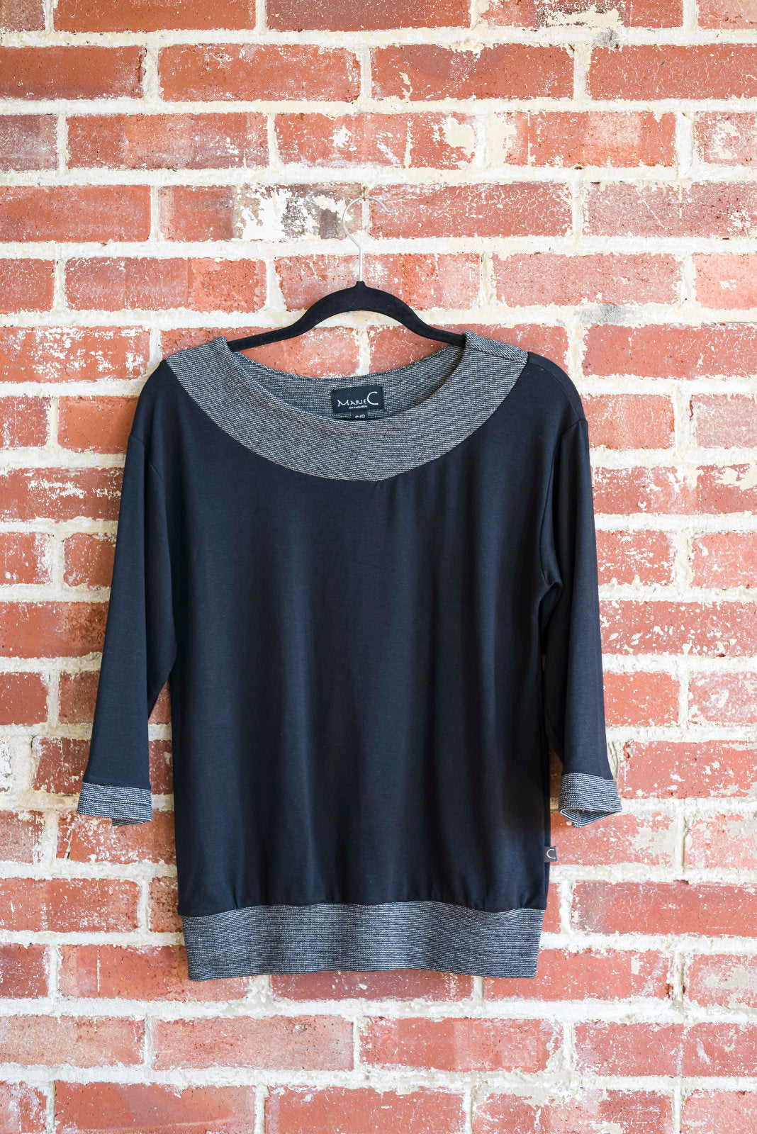 The Jaja Top by Marie C in Black is shown hanging against a brick wall. It's a sweatshirt with contrasting fabric bands at the wide neck, 7/8 sleeves, and hem.