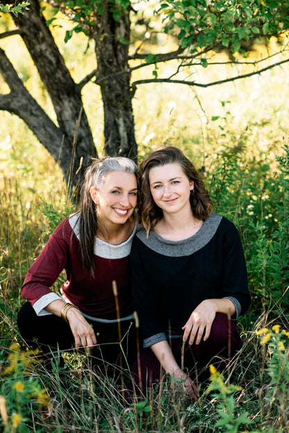 Two women wearing the Jaja Top by Marie C, one in Black and one in Wine. It's a sweatshirt with contrasting fabric bands at the wide neck, 7/8 sleeves, and hem. They are sitting in front of a tree. 