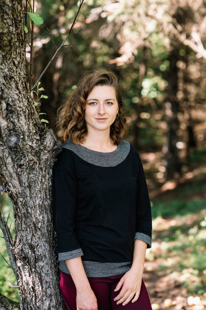 A woman wearing the Jaja Top by Marie C in Black. It's a sweatshirt with contrasting fabric bands at the wide neck, 7/8 sleeves, and hem. She is standing in front of a tree. 