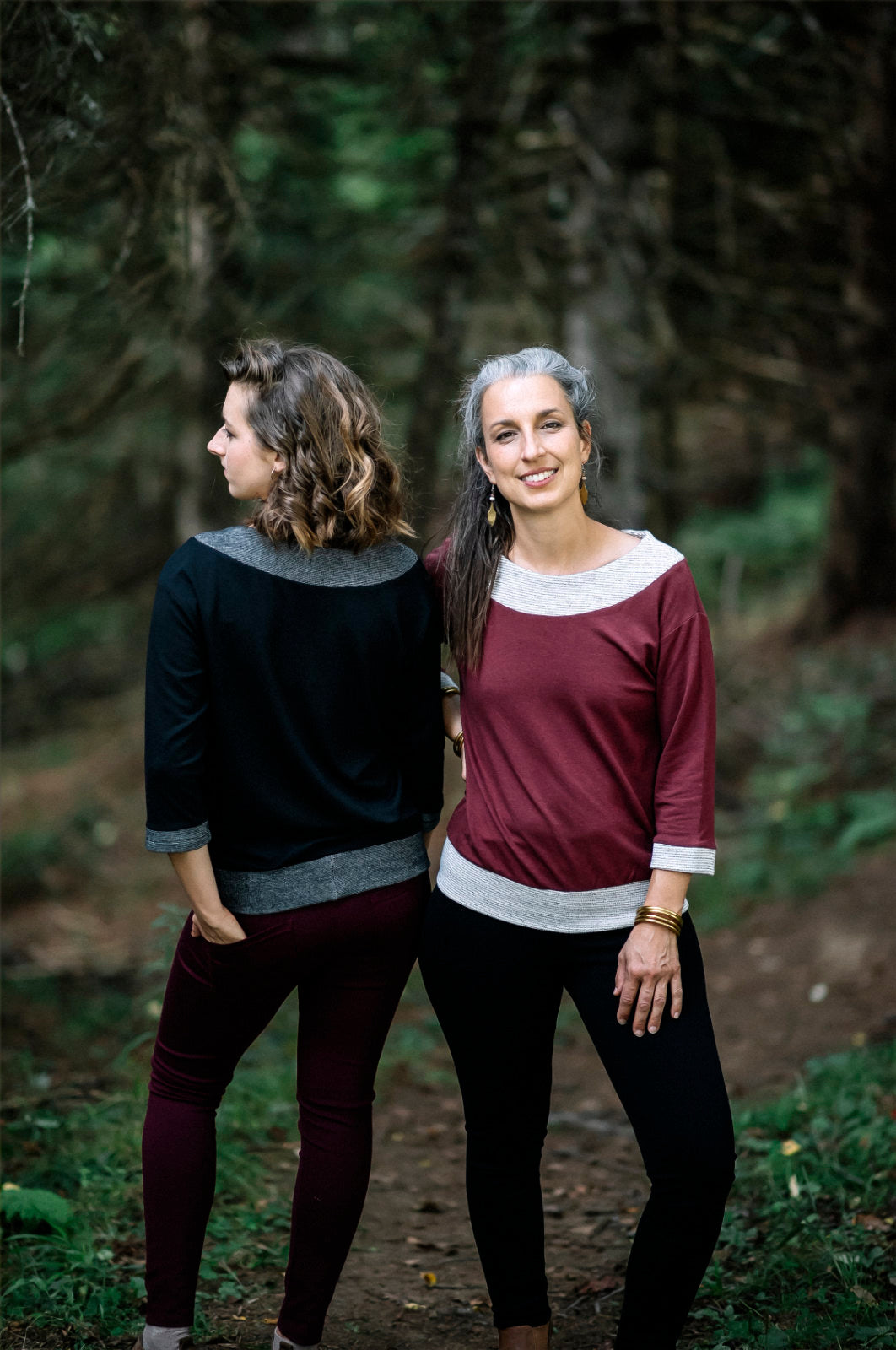 Two women wearing the Jaja Top by Marie C, one in Black and one in Wine. It's a sweatshirt with contrasting fabric bands at the wide neck, 7/8 sleeves, and hem. They are standing on a forest path.