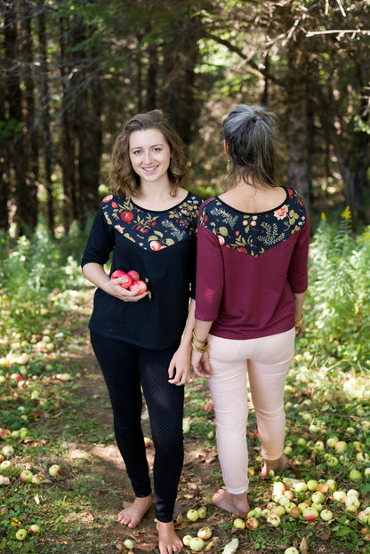 Two women wearing the Manzana Top by Marie C, one in Black and one in Wine. It's a reversible top with autumn print fabric along the neck on one side, and 3/4 sleeves. They are standing in in an apple orchard. 