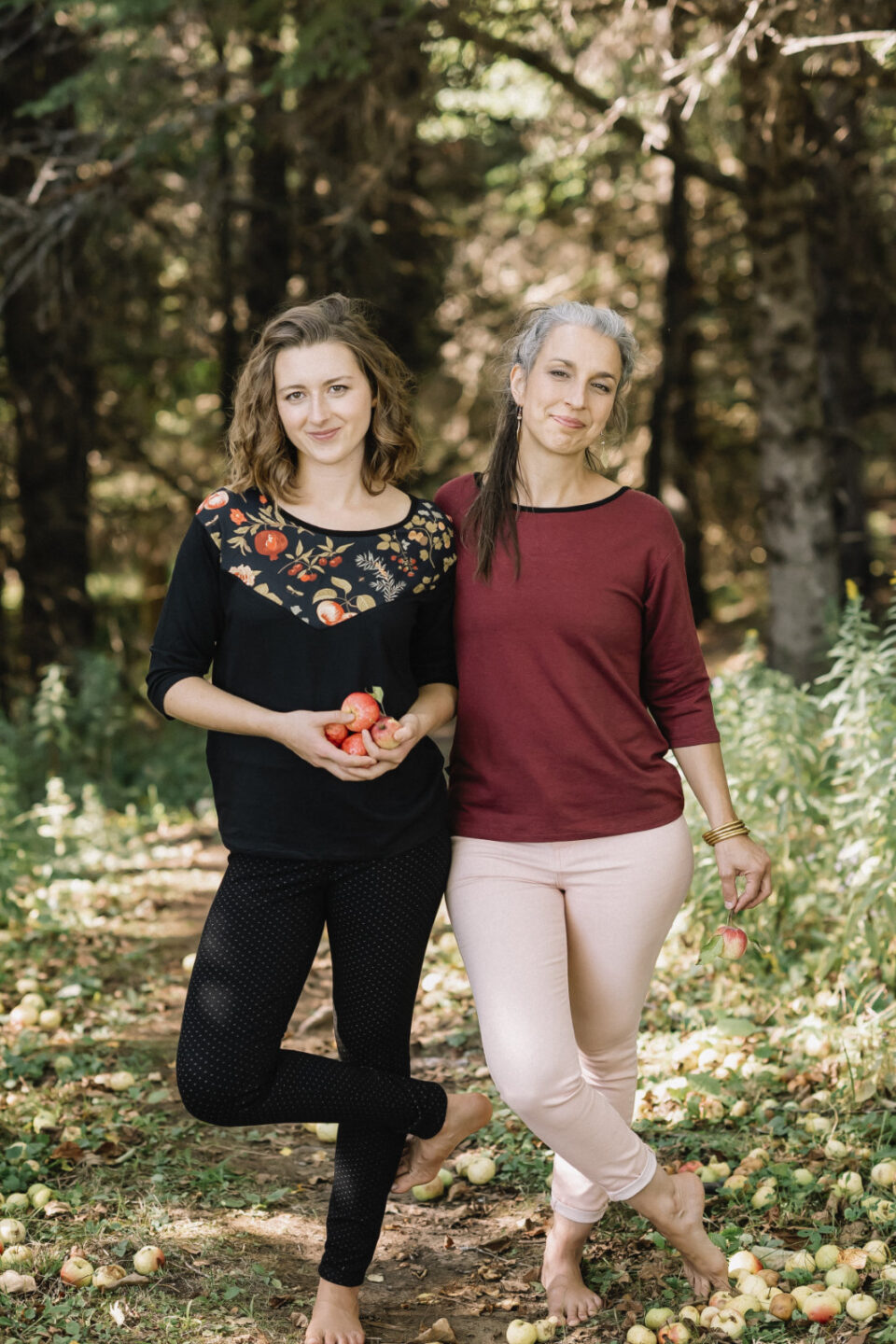 Two women wearing the Manzana Top by Marie C, one in Black and one in Wine. It's a reversible top with autumn print fabric along the neck on one side, and 3/4 sleeves. They are standing in in an apple orchard. 