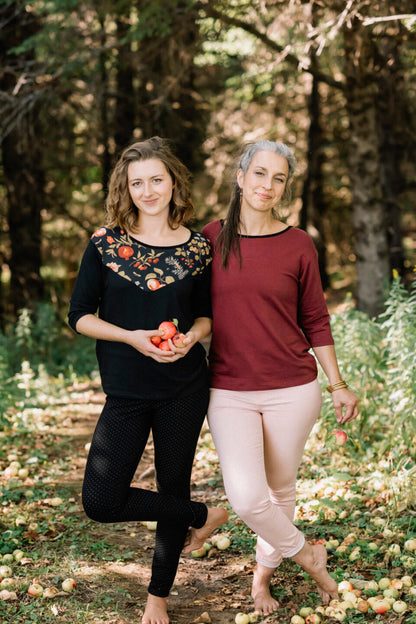 Two women wearing the Manzana Top by Marie C, one in Black and one in Wine. It's a reversible top with autumn print fabric along the neck on one side, and 3/4 sleeves. They are standing in in an apple orchard. 