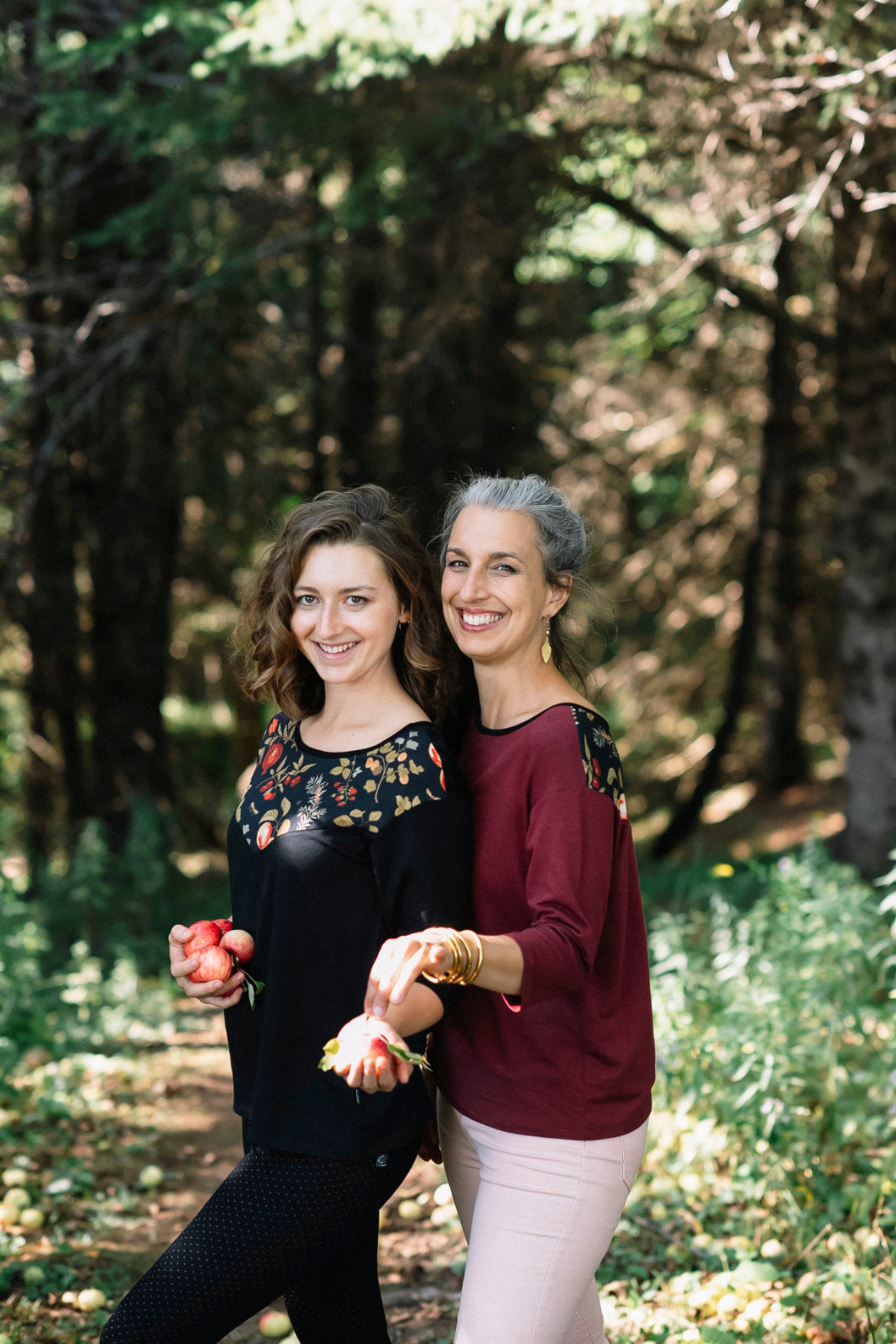 Two women wearing the Manzana Top by Marie C, one in Black and one in Wine. It's a reversible top with autumn print fabric along the neck on one side, and 3/4 sleeves. They are standing in in an apple orchard. 