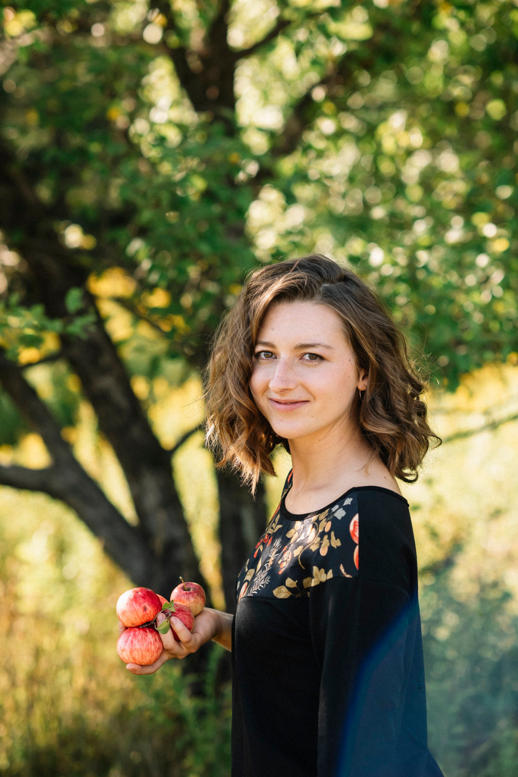 Side view of a woman wearing the Manzana Top by Marie C in Black. It's a reversible top with autumn print fabric along the neck on one side, and 3/4 sleeves. She is  standing in in an apple orchard. 