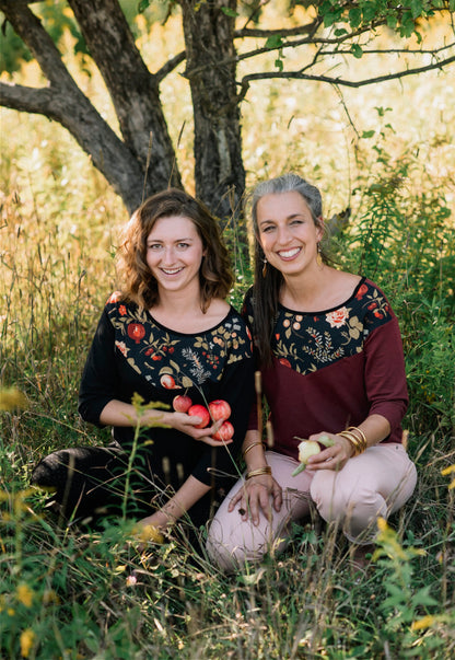 Two women wearing the Manzana Top by Marie C, one in Black and one in Wine. It's a reversible top with autumn print fabric along the neck on one side, and 3/4 sleeves. They are standing in in an apple orchard. 