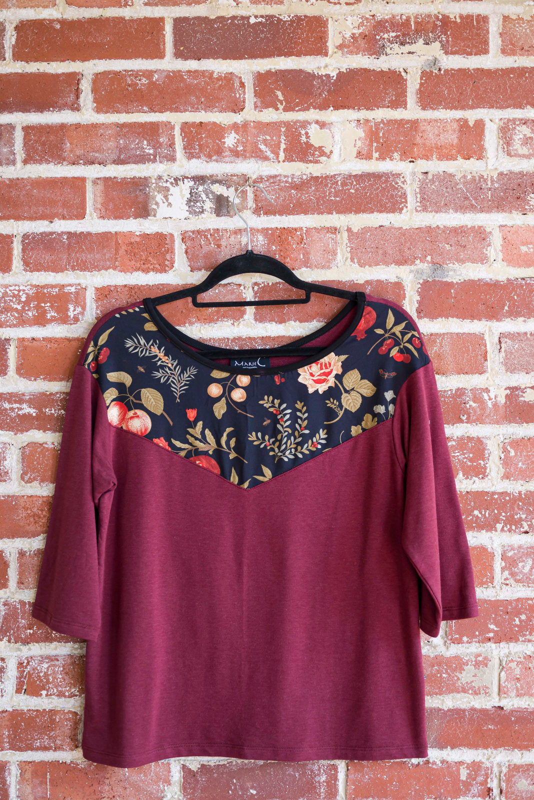 The Manzana Top by Marie C in Wine is shown hanging against a brick wall. It's a reversible top with autumn print fabric along the neck on one side, and 3/4 sleeves. 