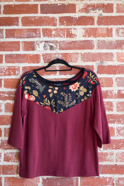 The Manzana Top by Marie C in Wine is shown hanging against a brick wall. It's a reversible top with autumn print fabric along the neck on one side, and 3/4 sleeves. 