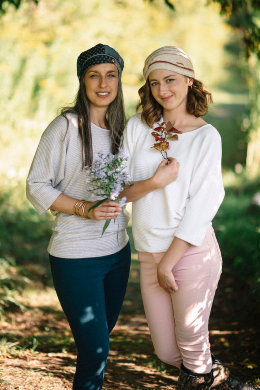 Two women wearing the Perla Top by Marie C,  one in Ivory and one in Stripes, with a wide round neck, 3/4 batwing sleeves, and fitted waist in ribbed knit fabric. They are standing outside. 