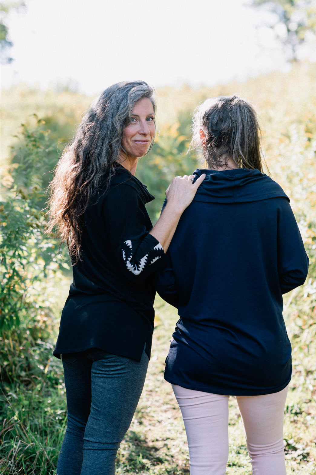 Back view of two women wearing the Tunic Java by Marie C, one in Black and one in Navy, featuring a cowl collar, 7/8 batwing sleeves with contrast fabric, and a hi-low hemline with a slit detail. They are standing in a forest. 