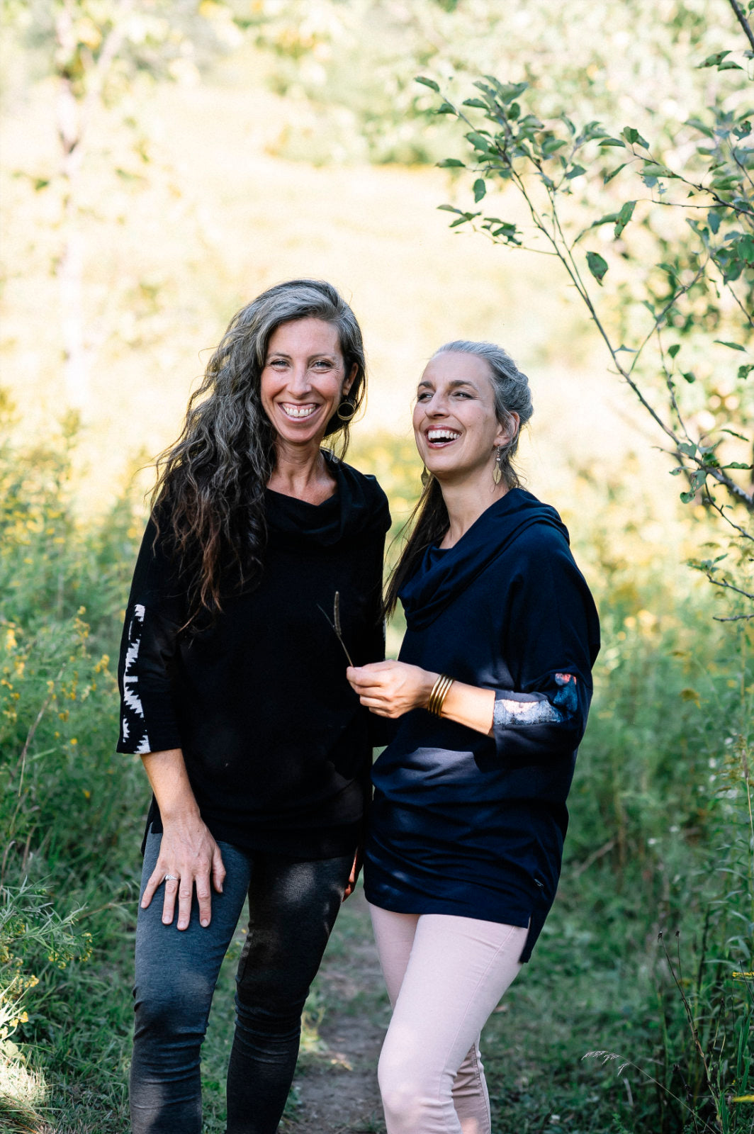 Two women wearing the Tunic Java by Marie C, one in Black and one in Navy, featuring a cowl collar, 7/8 batwing sleeves with contrast fabric, and a hi-low hemline with a slit detail. They are standing in a forest. 