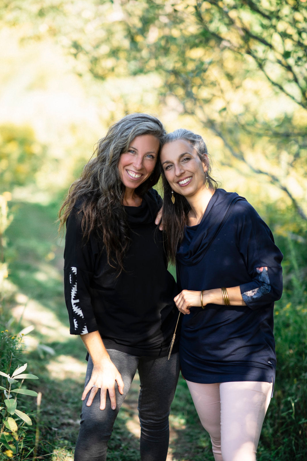 Two women wearing the Tunic Java by Marie C, one in Black and one in Navy, featuring a cowl collar, 7/8 batwing sleeves with contrast fabric, and a hi-low hemline with a slit detail. They are standing in a forest. 