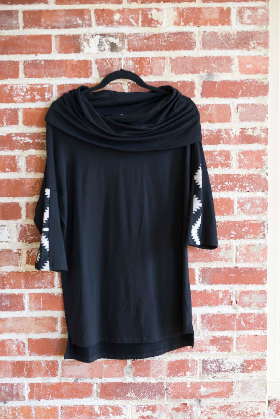 The tunic Java by Marie C in Black is shown hanging against a brick wall. It features a cowl collar, 7/8 batwing sleeves with contrast fabric, and a hi-low hemline with a slit detail. 