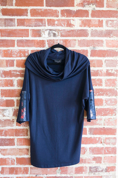 The tunic Java by Marie C in Navy is shown hanging against a brick wall. It features a cowl collar, 7/8 batwing sleeves with contrast fabric, and a hi-low hemline with a slit detail. 