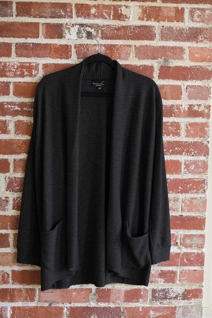 The Margo Cardigan by Marie C in Black, a long open cardigan  in ribbed knit with rounded patch pockets and dropped sleeves, is shown hanging in front of a brick wall. 
