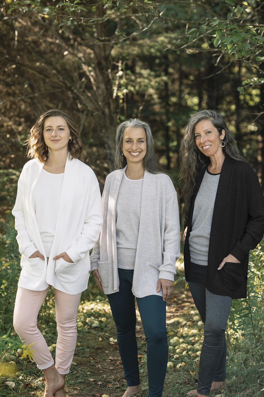 Three women wearing the Margo Cardigan by Marie C in Ivory, stripes, and black, a long open cardigan  in ribbed knit with rounded patch pockets and dropped sleeves. They are wearing them with jeans and standing in a forest.
