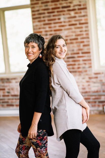 Two women standing back to back, wearing the Juliana Tunic by Marie C in Black and Sand, with a crossover neck, 3/4 sleeves, hi-low hemline, and ribbed fabric. They are standing in front of a brick wall. 