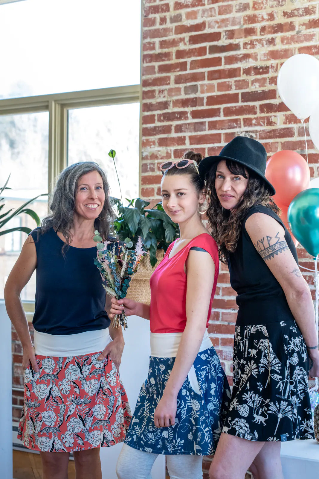 Three women wearing the Sol Top from Marie C, a reversible cap sleeve tee. From left to right they are wearing it in Navy, Coral, and Black. They are all wearing skirts and are standing at a party. 