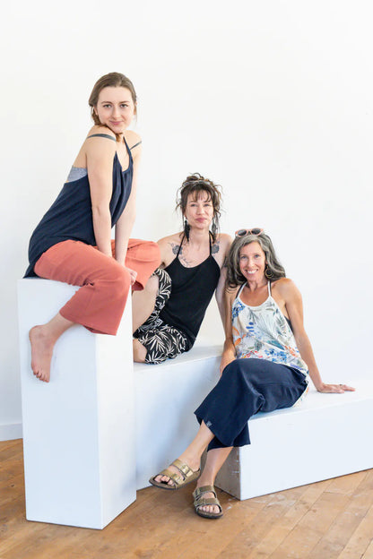 Three women wearing the Verano Pants by Marie C in various colours, they are wearing them with tank tops and sitting on white blocks in front of a white wall. 