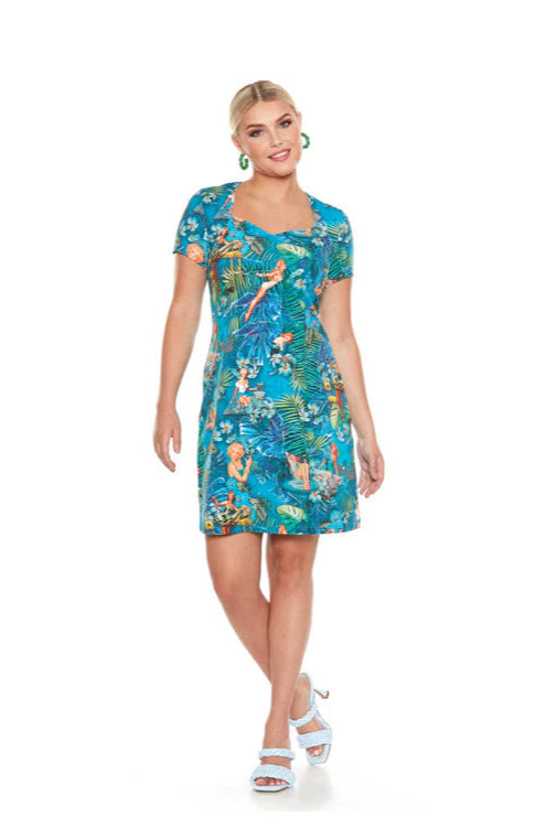 A woman wearing the Marilyn Dress by Yul Voy, featuring a vintage inspired tropical print with pin-up girls on it, a sweetheart neckline, short sleeves, and a sheath shape that falls above the knee. 