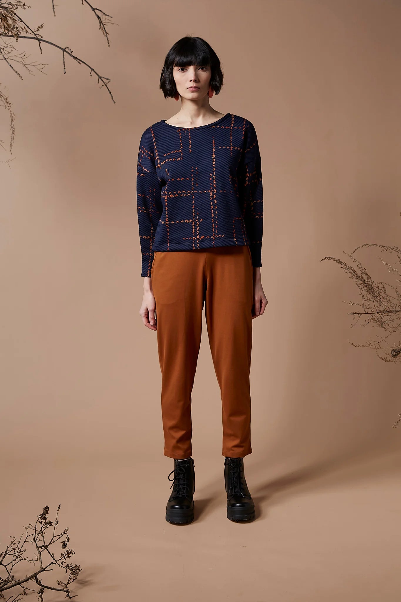 A woman wearing the Jackson Top by Melow in indigo with orange pants, standing in front of a peach background 