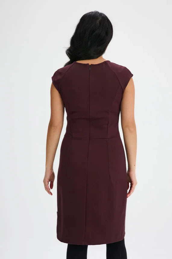 Back view of a woman wearing the Ivry Dress by Message Factory in Burgundy, a knee-length dress mad from a Lenzing Ecovero viscose blend, featuring a round neck and cap sleeves with princess seams and a defined waist. She is standing in front of a white background. 