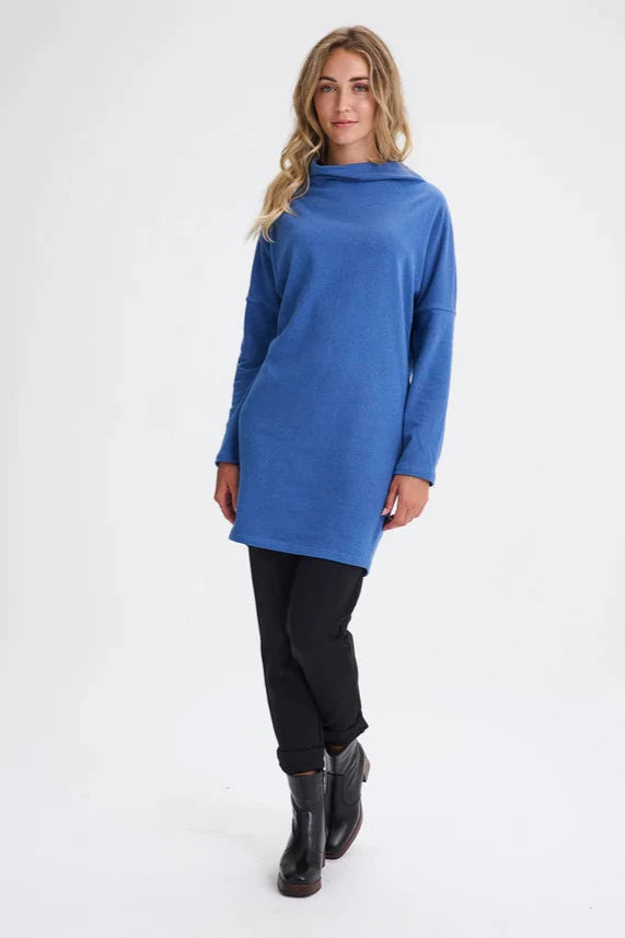 A woman wearing the Abilene Dress by Message Factory in Blue, with a large cowl neck, bat wing sleeves, and a mid-thigh length, all made from recycled cotton.
She is wearing it with slim black pants and standing in front of a white background. 