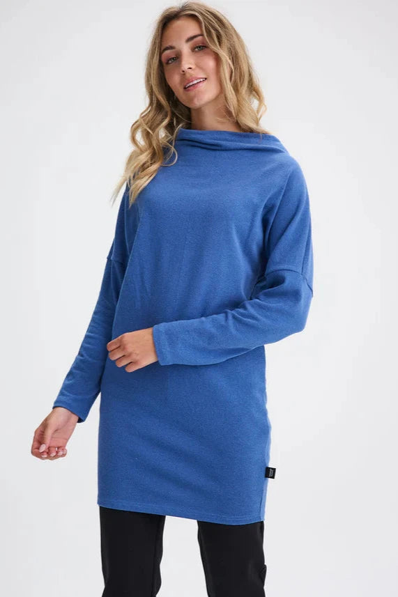 A woman wearing the Abilene Dress by Message Factory in Blue, with a large cowl neck, bat wing sleeves, and a mid-thigh length, all made from recycled cotton.
She is wearing it with slim black pants and standing in front of a white background. 