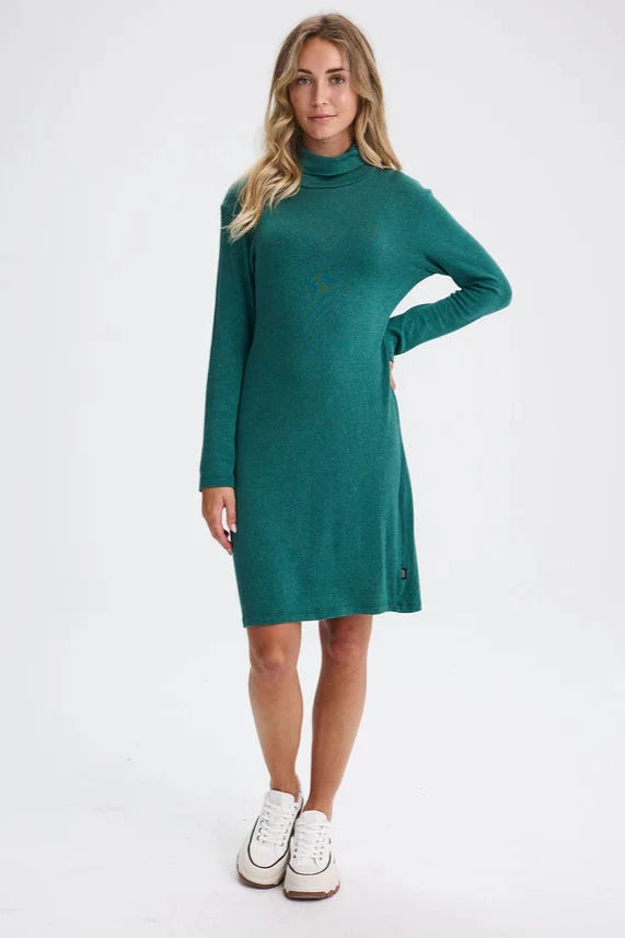 A woman wearing the Betula Dress by Message Factory in Evergreen, a knit turtleneck dress with long sleeves and a knee-length hem. She is standing in front of a beige background. 