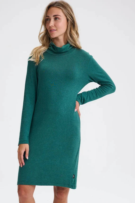 A woman wearing the Betula Dress by Message Factory in Evergreen, a knit turtleneck dress with long sleeves and a knee-length hem. She is standing in front of a beige background. 