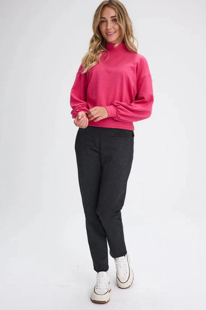 A woman wearing the Fermont Sweatshirt by Message Factory in Fuchsia, made from bamboo and cotton with a high neck, long sleeves with a puff detail at the cuffs, and fitted band at the bottom. She is wearing it with grey pants and is standing in front of a white background. 