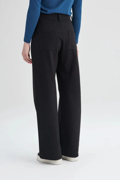 Back waist down view of a woman wearing the Hatley Pants by Message Factory in Black, a wide leg pant with a button fly and two pockets, made from Lenzing Ecovero viscose, with a blue top. She is standing in front of a white background. 