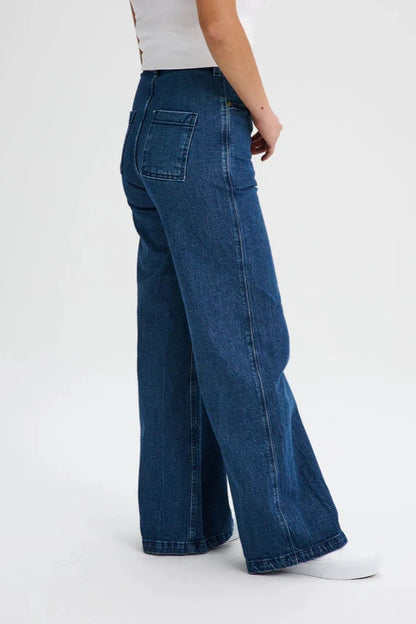 Back waist down view of a woman wearing the Hochelaga Flared Leg Denim Pants by Message Factory, a wide leg jean made from organic cotton, with a white tee. She is standing in front of a white background. 