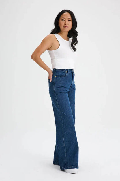 A  woman wearing the Hochelaga Flared Leg Denim Pants by Message Factory, a wide leg jean made from organic cotton, with a white tank. She is standing in front of a white background. 