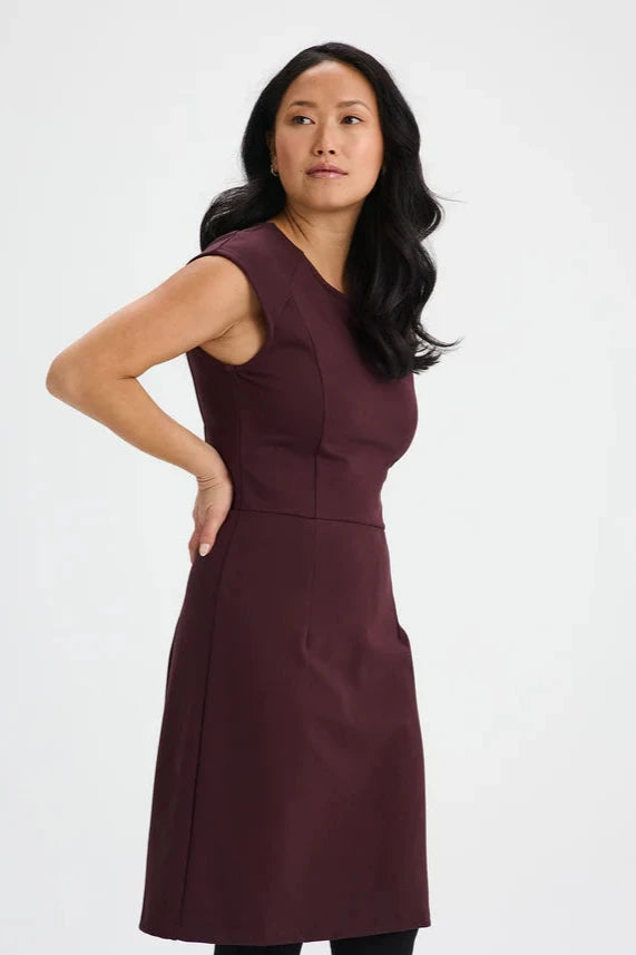 Side view of a woman wearing the Ivry Dress by Message Factory in Burgundy, a knee-length dress mad from a Lenzing Ecovero viscose blend, featuring a round neck and cap sleeves with princess seams and a defined waist. She is standing in front of a white background. 