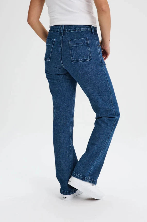 Back waist down view of a woman wearing the Marianne Straight Leg Denim Pants by Message Factory, featuring a straight leg, a 31 inch inseam, and organic cotton fabric, with a white tank. She is standing in front of a white background. 