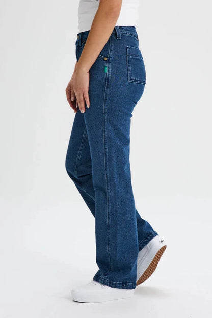 Side waist down view of a woman wearing the Marianne Straight Leg Denim Pants by Message Factory, featuring a straight leg, a 31 inch inseam, and organic cotton fabric, with a white tank. She is standing in front of a white background. 