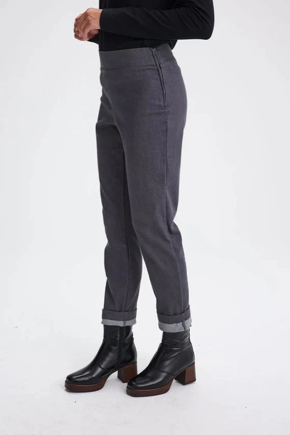 Waist down side view of a woman wearing the Potton Pants by Message Factory in Gray Denim,  slim cut pants in stretchy denim with a wide waistband and a side zipper, made from recycled cotton and polyester. She's standing in front of a white background. 